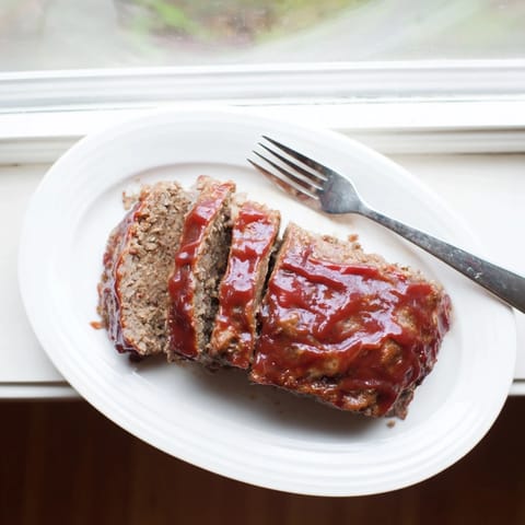 Slices of juicy classic Meatloaf topped with sticky ketchup glaze, served alongside creamy mashed potatoes and buttered green beans on a dinner plate.